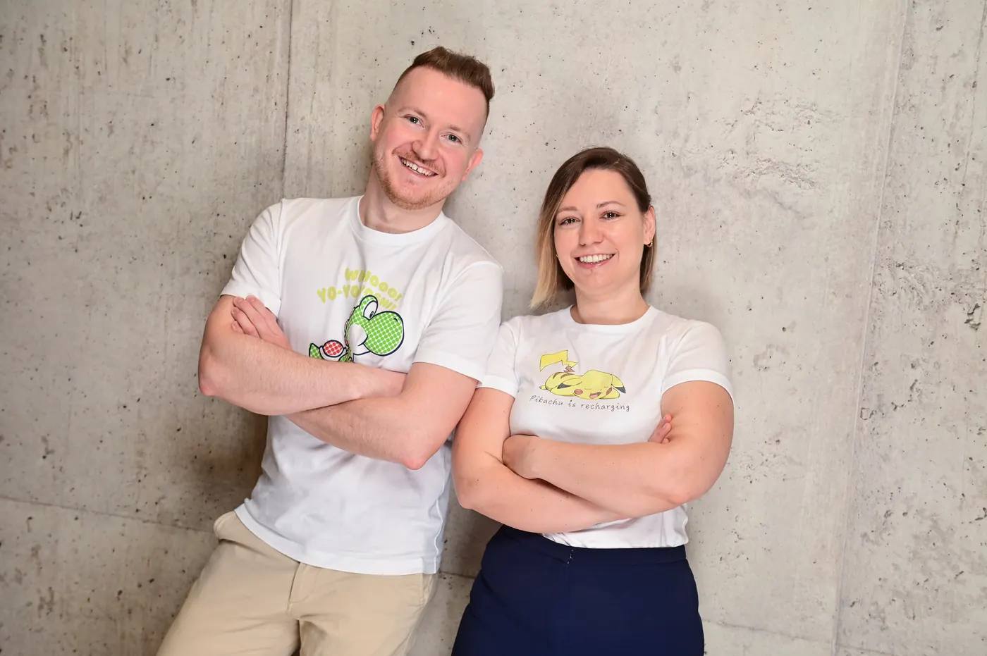 Simona und Jürgen lehnen lächelnd mit verschränkten Armen an einer Betonwand, beide in weißen T-Shirts mit Spielefiguren